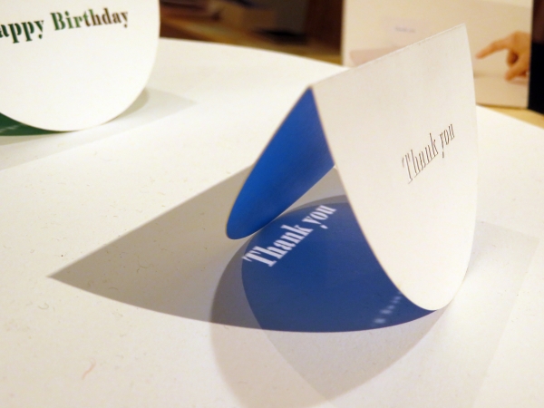 お祝いや感謝の言葉をレーザー加工で表現したグリーティングカードは、平らな面に置いて、指で押すと揺れ動き、光に当てるとメッセージが影となり机上に映し出される。 大成紙器製作所 